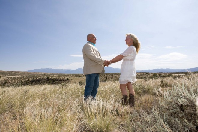 Jeff and Michelle hold hands in the long grass with Taos Mountains in the background