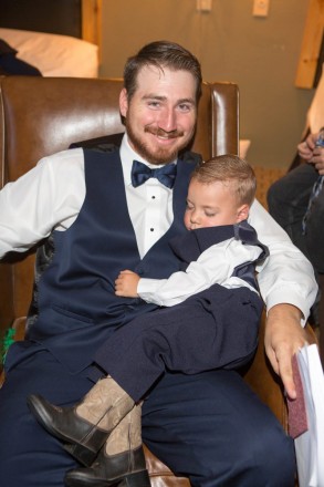 George holds his sleeping two-year-old son and ring bearer who is wearing cowboy boots with his tuxedo.