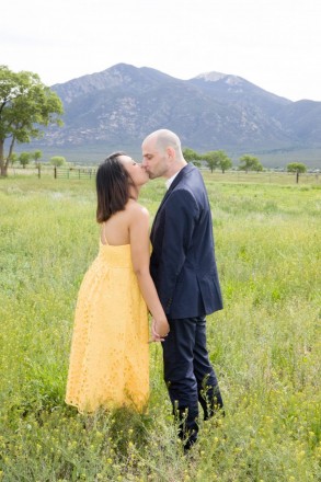 Kath and Brian share a kiss in front of magical Taos Mountain in a green meadow with yellow wildflowers