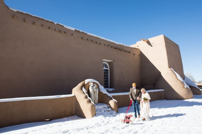 Bride and groom walk past San Francisco de Asis church in Rachos de Taos with blanket of snow...