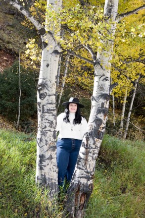 Katy, in jeans and a white sweater, smiles between golden aspen trees