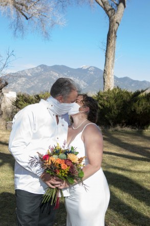 Bride and groom, both in white, in front of Taos Mountain, kissing with masks on.