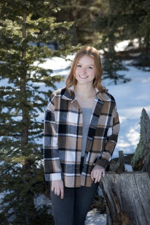 Senior in flannel shirt and jeans stands with pine tree for snowy senior shoot
