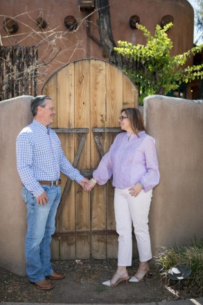 Couple pose for a photo with a latilla fence,  adobe home with vigas, and a wooden door.