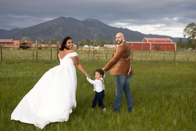 Bride, groom, and ringbearer on wedding night with green meadow and red barn in Taos, NM