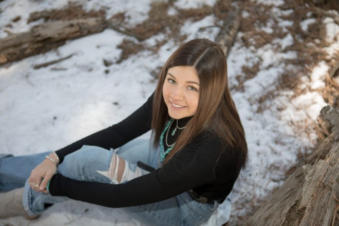 Sitting in the snow, high school senior smiles as she looks up at camera