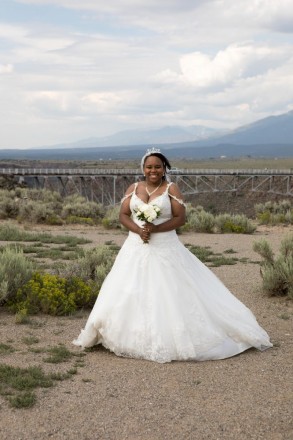 Cynthia smiles with bouquet of white roses in front of the Rio Grande gorge bridge